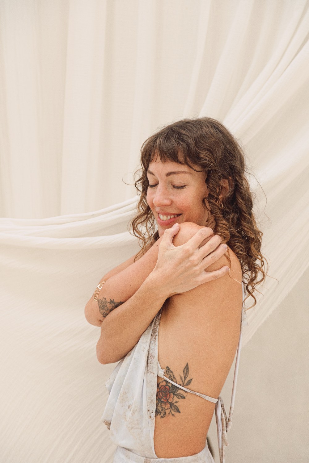 Ceci en autoabrazo, sonriendo con los ojos cerrados, fondo de tela crema.